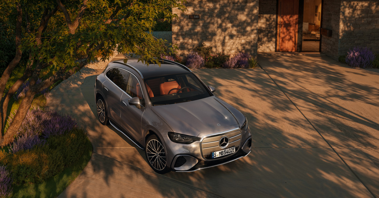 Der neue GLC. Silberner Mercedes-Benz SUV auf moderner Einfahrt vor einem zeitgenössischen Wohnhaus bei Sonnenaufgang oder Sonnenuntergang, umgeben von gepflegtem Garten mit lila Blumen und Bäumen.
