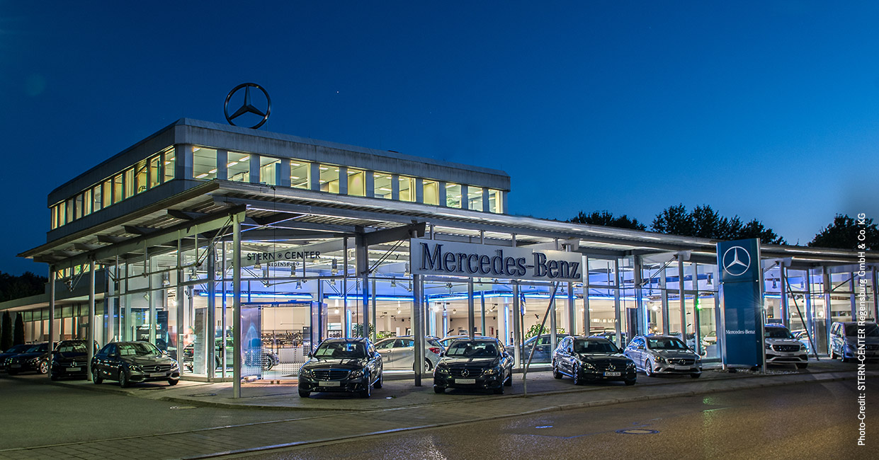 HIRSCHVOGEL Historie 2016 - Stern-Center Regensburg Dämmerungsaufnahme eines Mercedes-Benz Autohauses. Vor dem innen beleuchteten Gebäude stehen 7 Mercedes-Benz Fahrzeuge. Auf dem Dach ist ein Mercedes-Benz Stern angebracht. Auf der rechten Bildseite ist ein Mercedes-Benz Pylon zu erkennen.